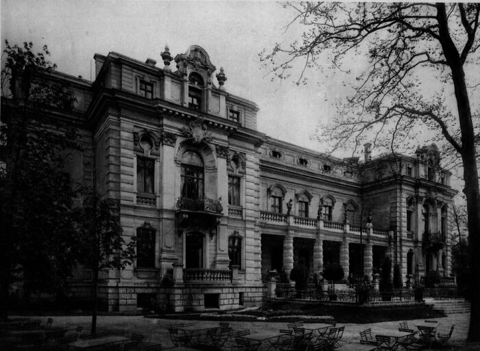 Black and white photo of a grand European building surrounded by trees and empty outdoor chairs, showing life in Europe.