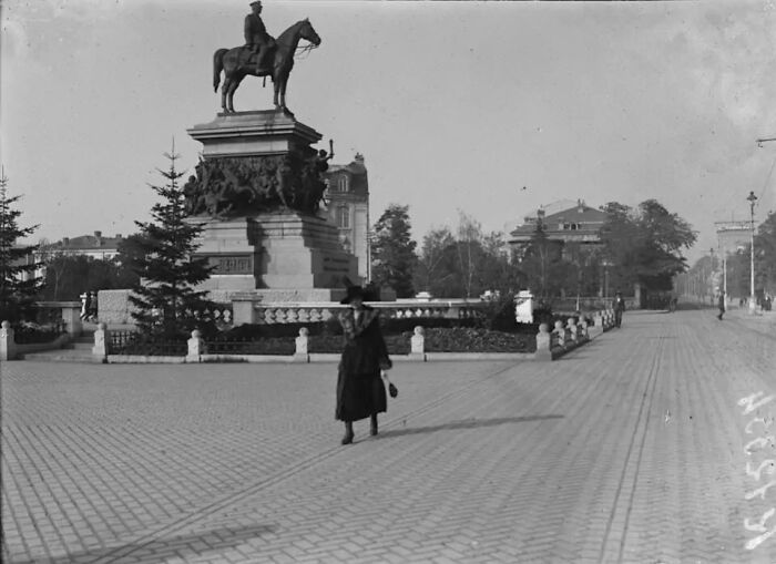Woman walking near a horse statue in a large paved city square, showing life in Europe 100 years ago.