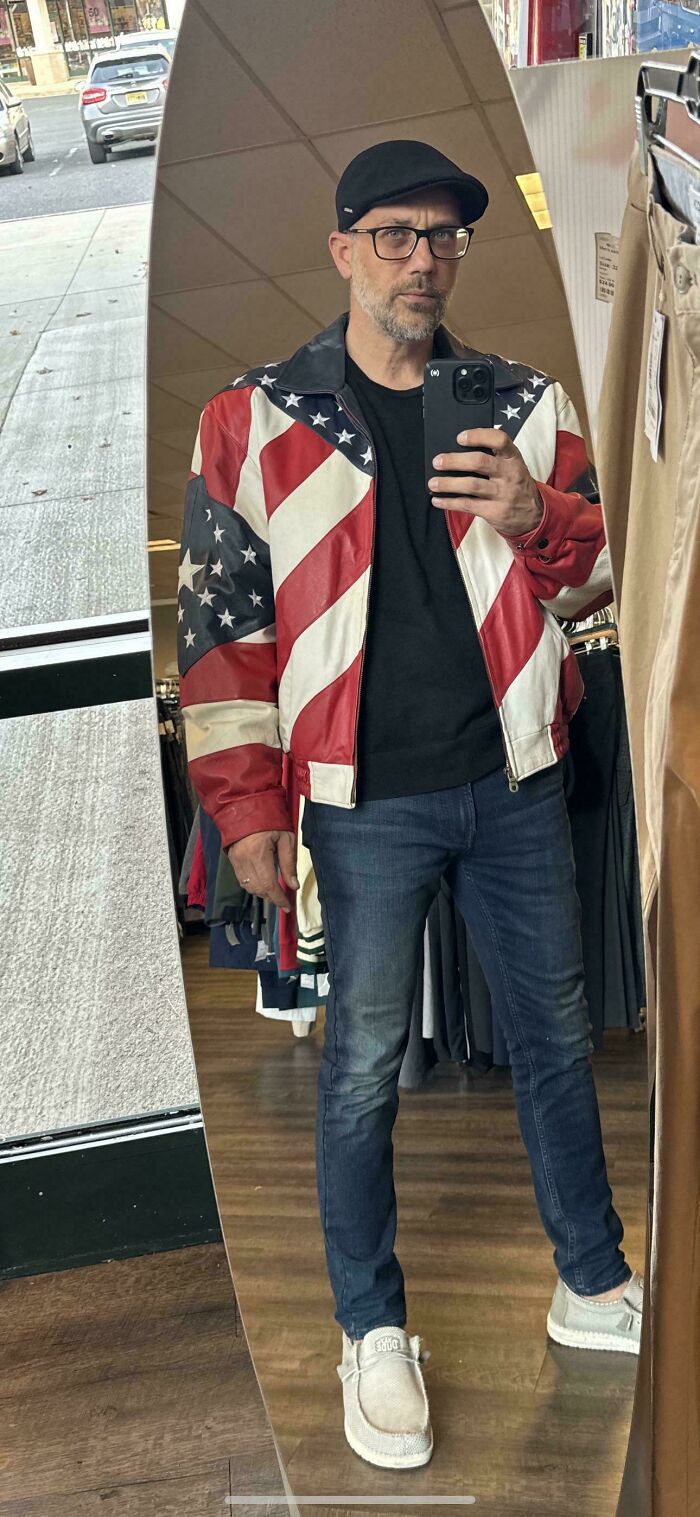 Man in mirror wearing a red, white, and blue jacket, found as a hidden gem at Goodwill, with jeans and a cap.