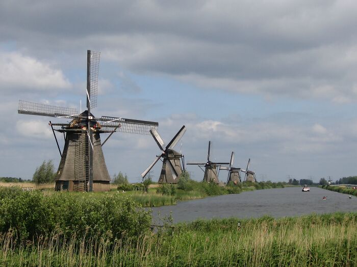 Four historic windmills by a river under a cloudy sky, showcasing architectural wonders not very well-known.