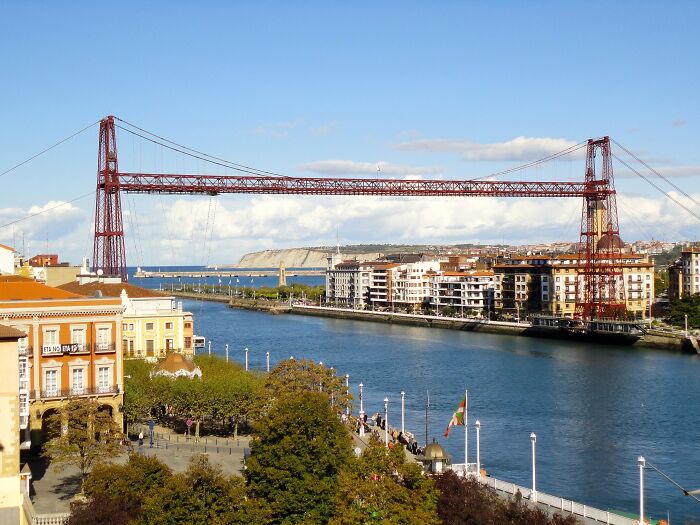 Architectural wonders: a stunning red iron bridge over a vibrant blue river, connecting bustling cityscapes under a clear sky.