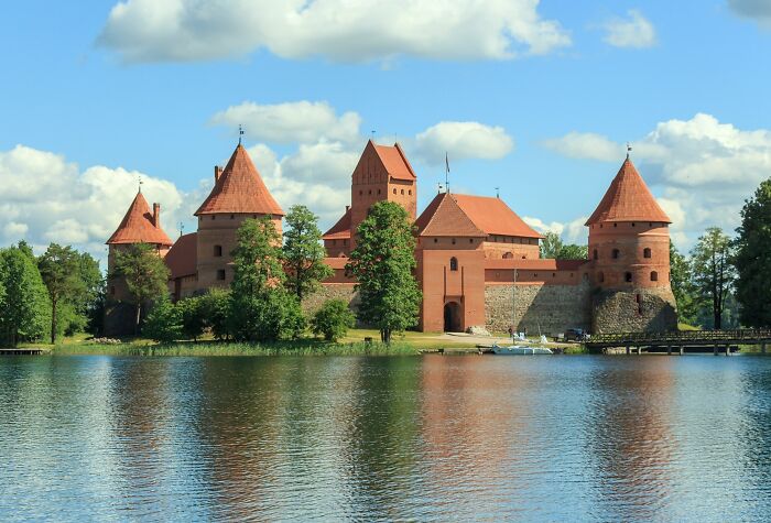 Red brick castle by a lake, surrounded by trees, exemplifying lesser-known architectural wonders.