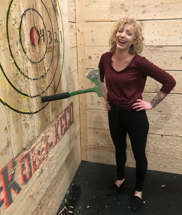 A woman smiling next to a mishap at an axe-throwing range, where an axe is lodged in the wall handle-first.