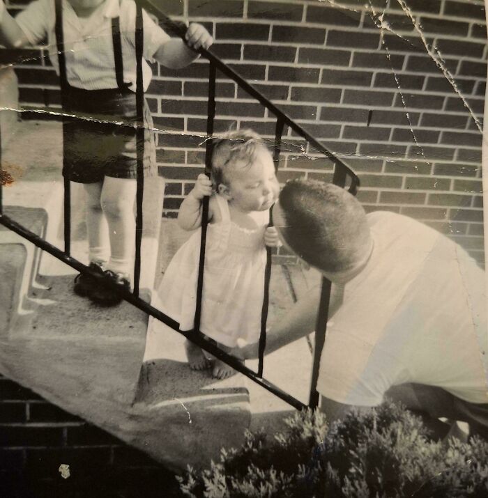Man kissing a baby through railing, with a child standing nearby; evokes nostalgia with this beautiful old pic.