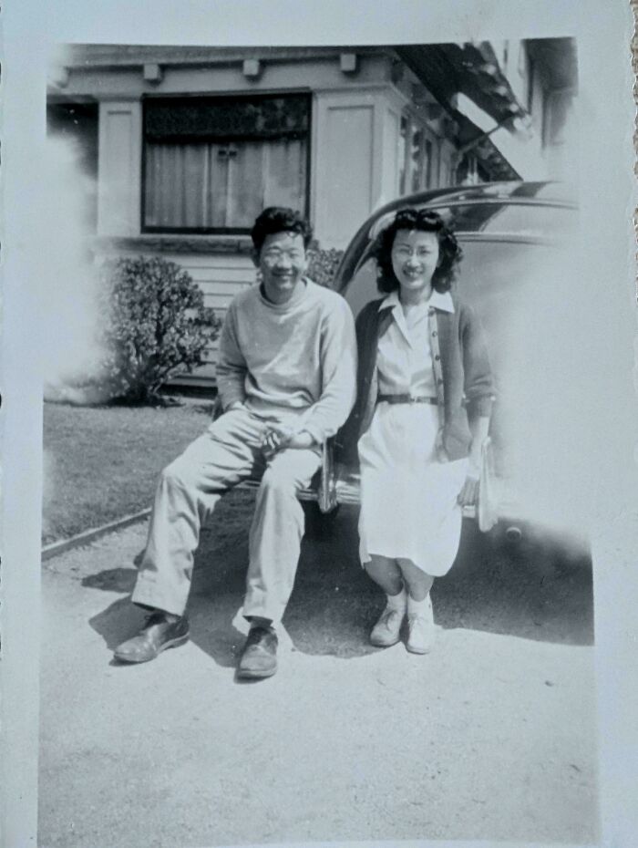 Pareja sonriente sentada junto a un auto antiguo frente a una casa, foto representativa de fotos de hace décadas.