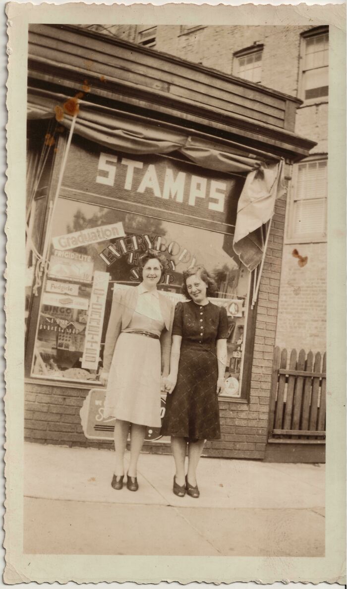 Dos mujeres de décadas pasadas posan sonrientes frente a una tienda de estampillas en una foto histórica.