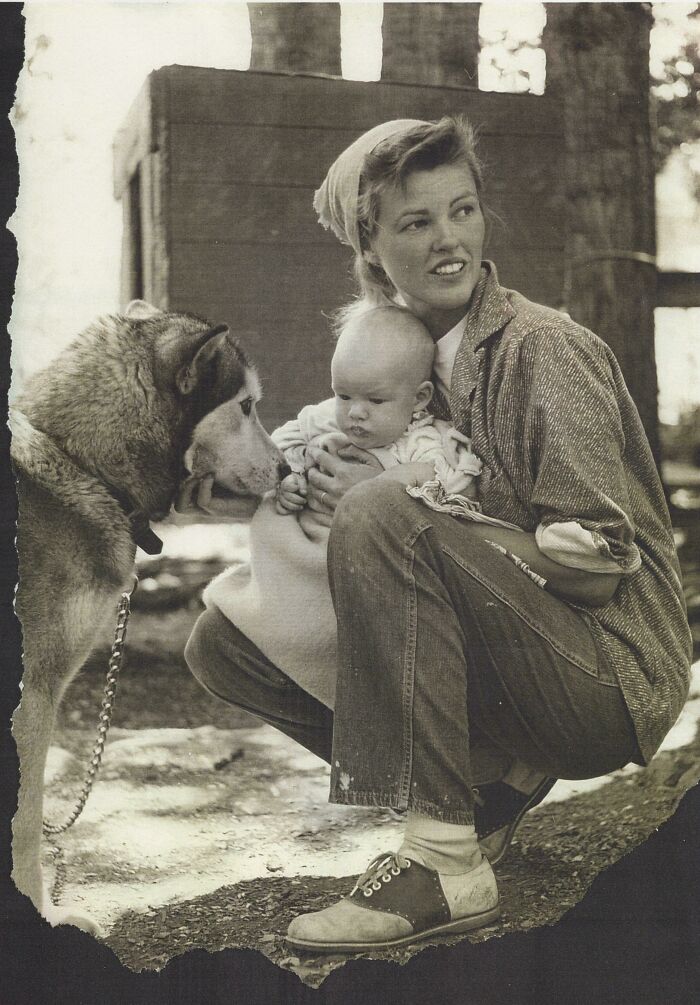 Mujer con bebé sosteniéndolo mientras un perro observa, foto antigua que refleja momentos elocuentes de décadas pasadas.