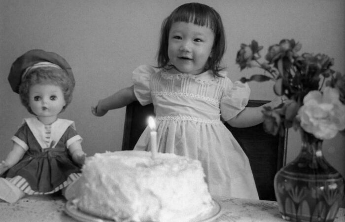 Vintage photo of a young child celebrating a birthday with a doll, cake, and flowers, bringing the past to life.