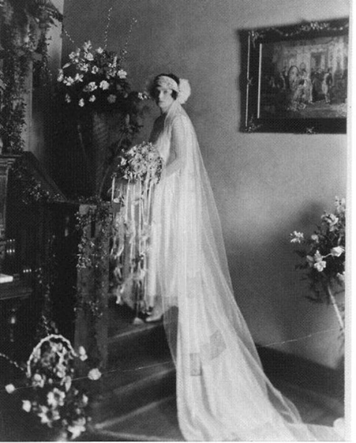 Vintage bride in elegant gown holding flowers, ascending stairs, surrounded by floral decor, evoking the beauty of the past.