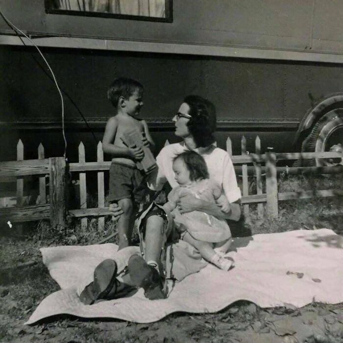 Vintage family picnic scene with a mother and two children sitting on a blanket outside near a wooden fence.
