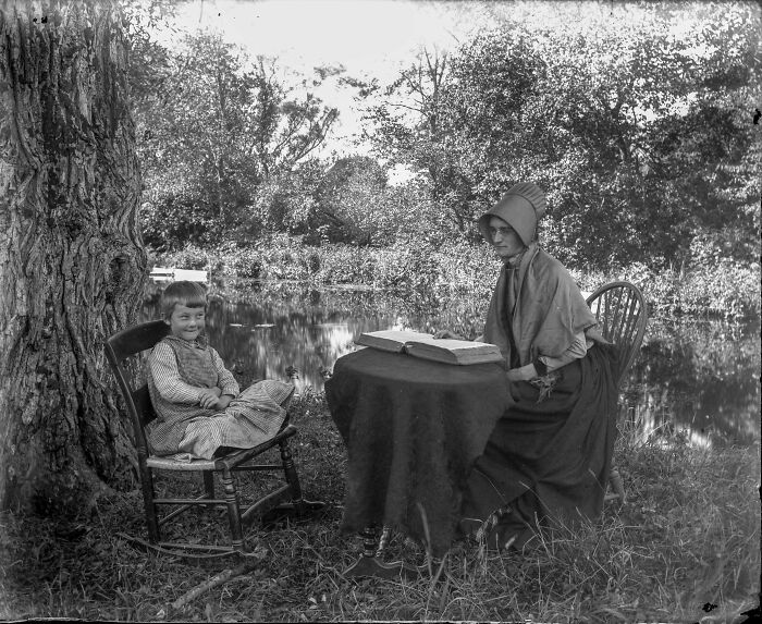 Old photo of a woman in a bonnet reading to a child by a river, surrounded by trees.