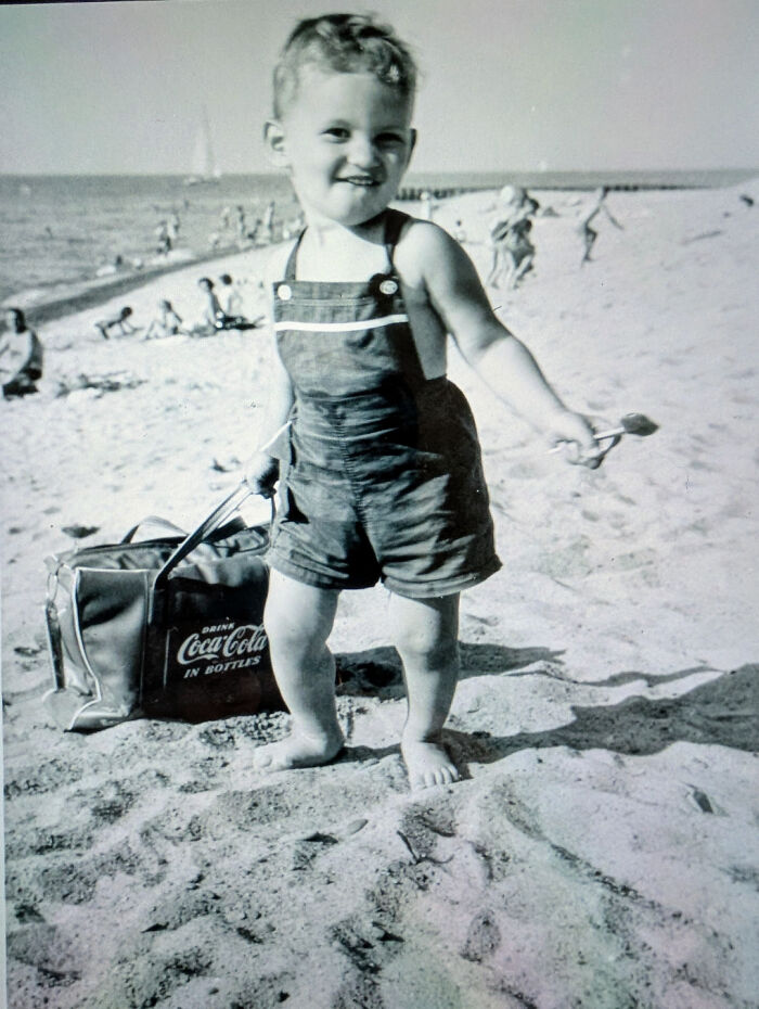 Young child on a vintage beach scene holding a shovel, with a classic Coca-Cola bag in the sand.