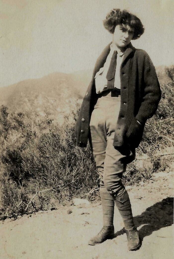 Old photo of a woman in early 20th-century attire, standing outdoors with mountains in the background.