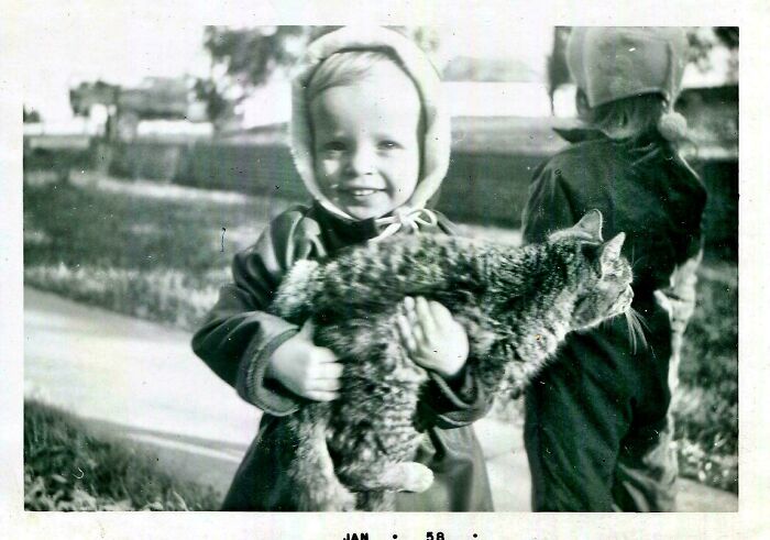 Child in winter clothing holding a cat, standing outdoors with another child nearby, capturing a beautiful moment from the past.