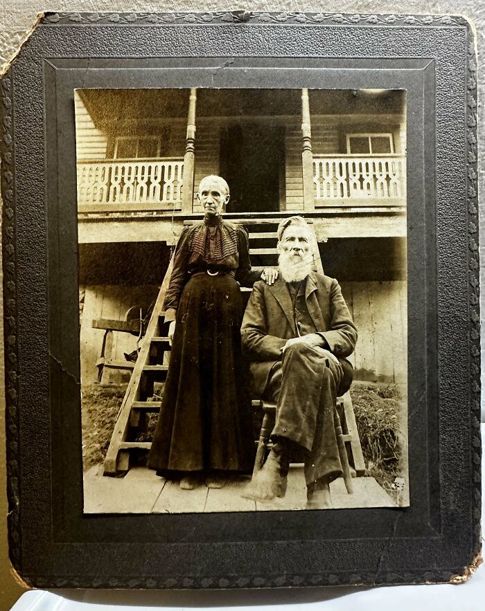 Elderly couple sitting and standing on porch, bringing the past back to life in a vintage photograph.