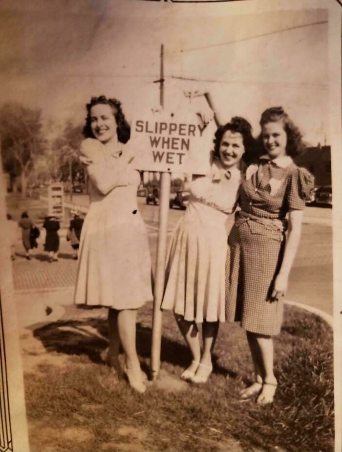 Three women in vintage dresses pose by a "Slippery When Wet" sign, capturing a beautiful moment from the past.