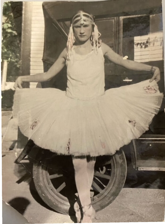 Old photo of a person in a ballet tutu and headpiece, standing by a vintage car, capturing historical charm.