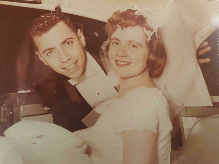 Vintage wedding photo of a smiling couple in a car, with the bride in a white gown and veil, and the groom in a tuxedo.