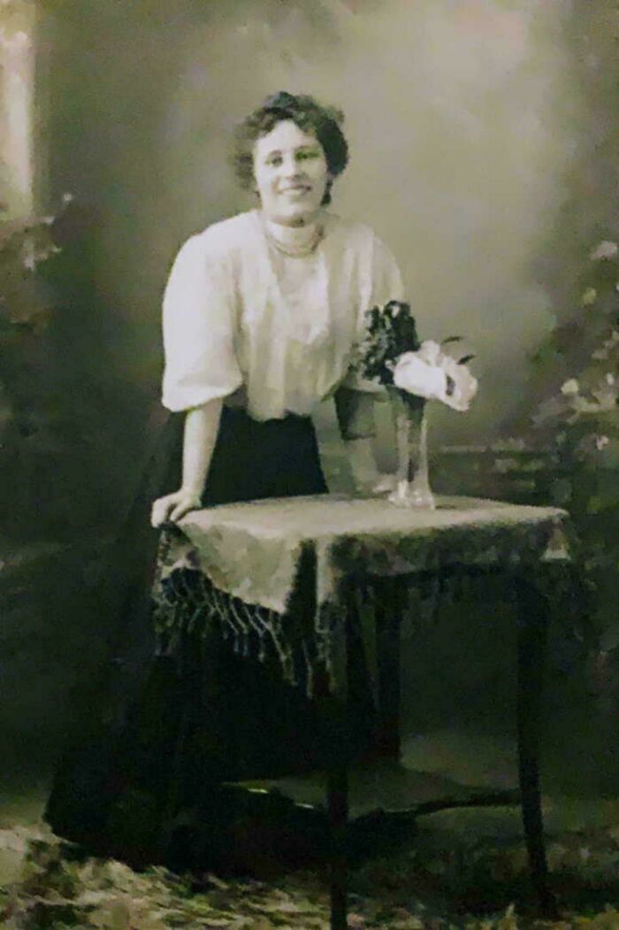 Mujer de época apoyada en mesa con mantel y flores, foto antigua representativa de fotos de hace décadas elocuentes.