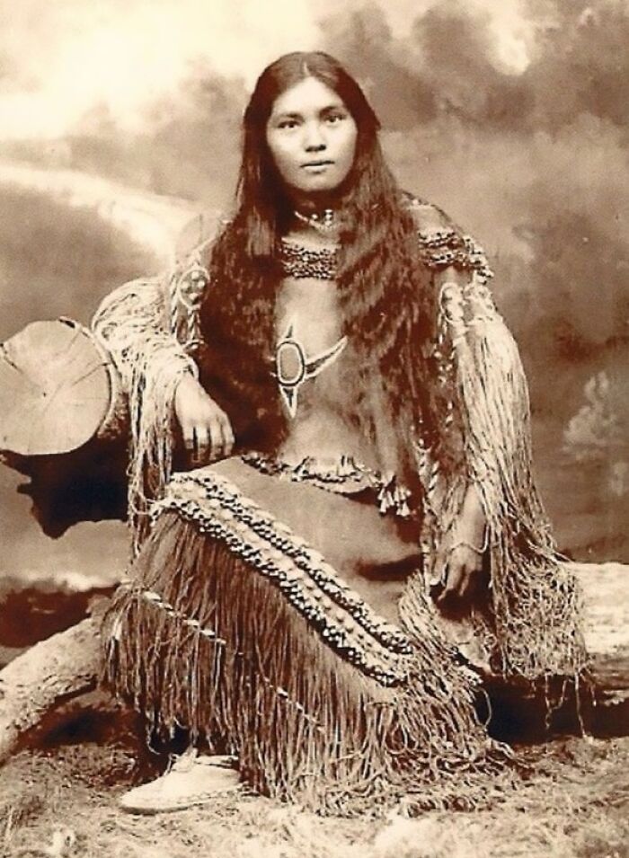 Native American woman in traditional dress sitting outdoors, captured in a haunting photo revealing real lives a century ago.