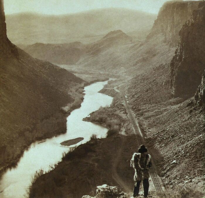 Native American man in traditional clothing overlooking a river valley and railroad tracks in a haunting historic landscape.