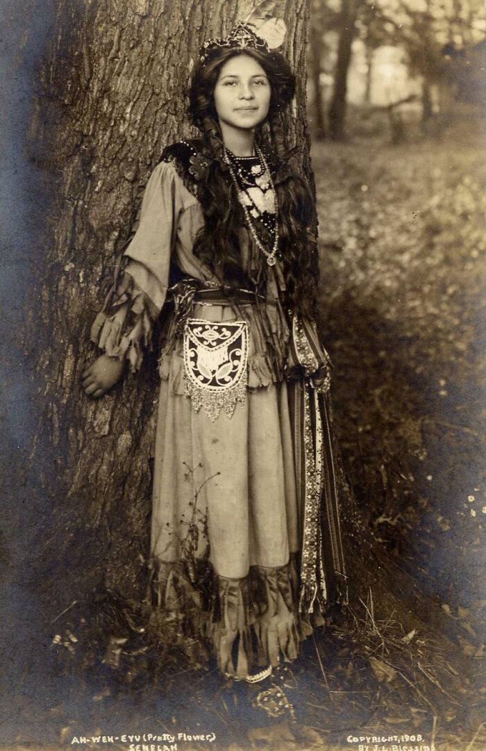 Native American woman in traditional dress standing by a tree in a haunting photo from a century ago.