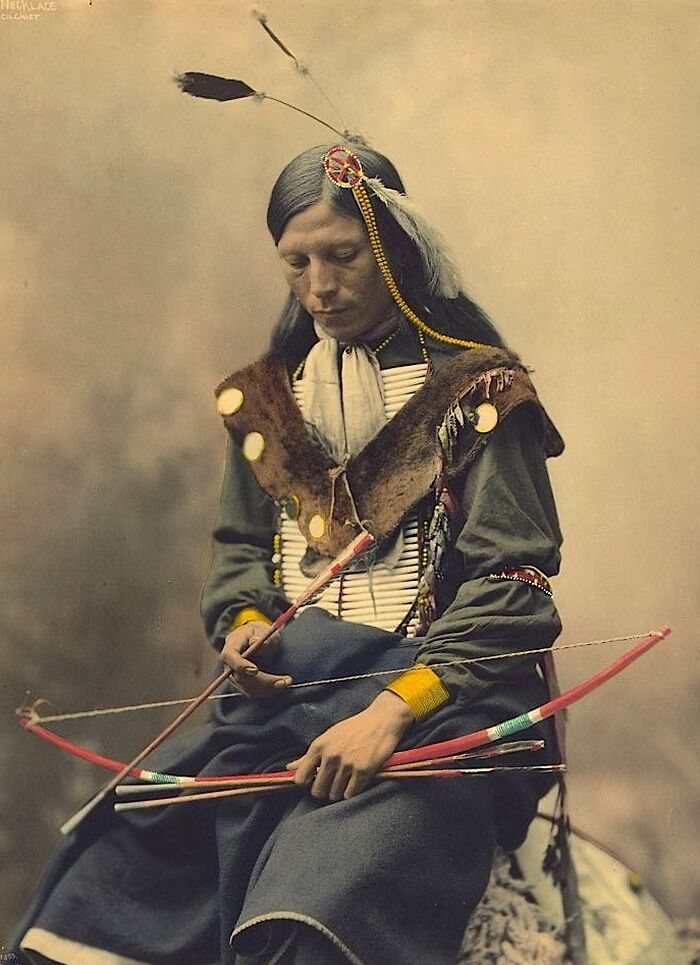 Native American man in traditional attire holding a bow and arrows, reflecting real lives of Native Americans a century ago.