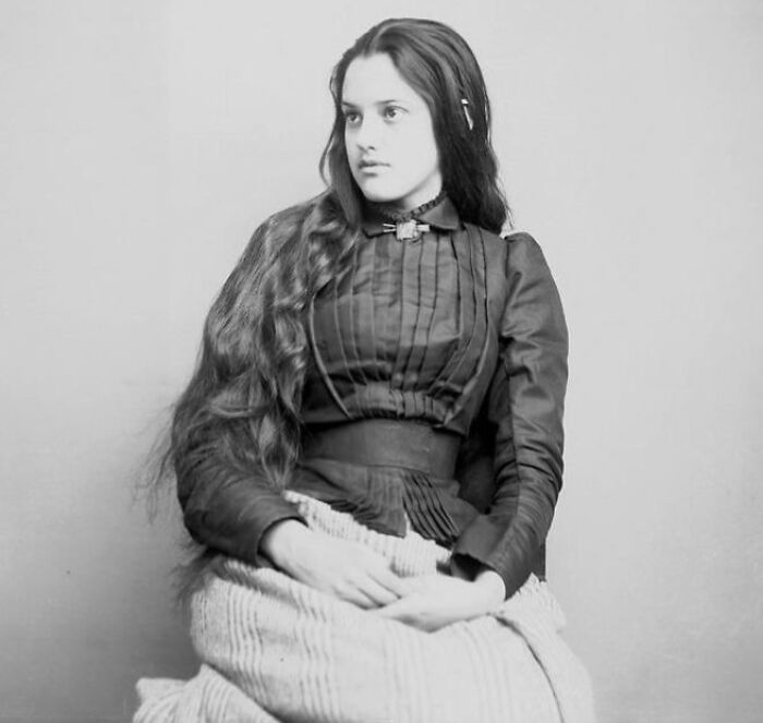 Native American woman from a century ago wearing traditional clothing and sitting with long hair against a plain backdrop