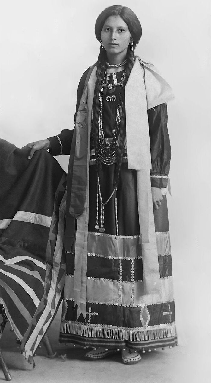 Native American woman in traditional attire with braided hair and beaded jewelry in a haunting photo from a century ago.