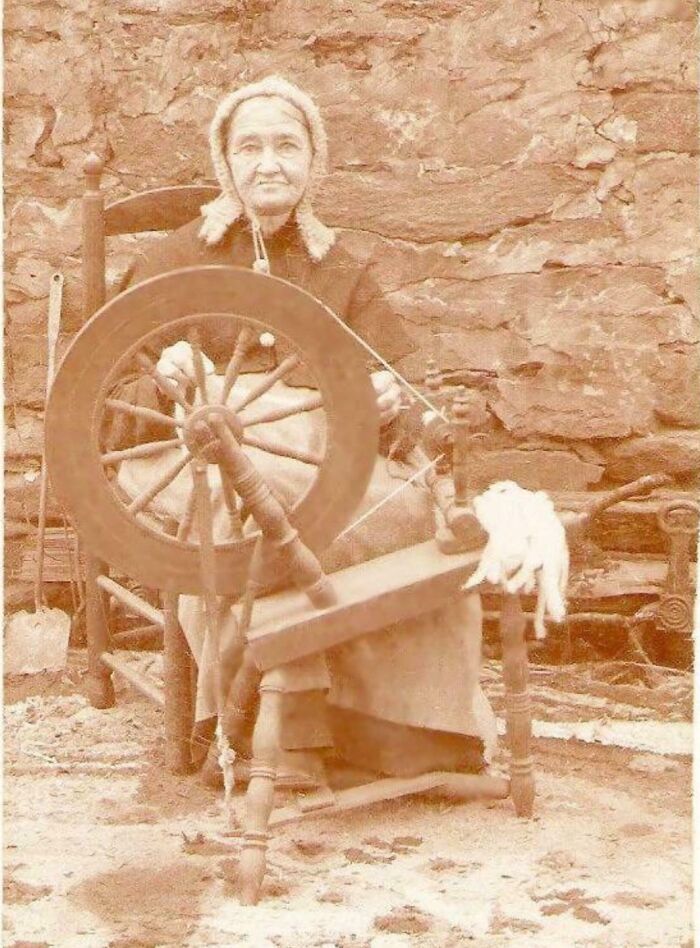 Mujer mayor hilando con rueca antigua, imagen histórica que refleja fotos de hace décadas con gran elocuencia visual.