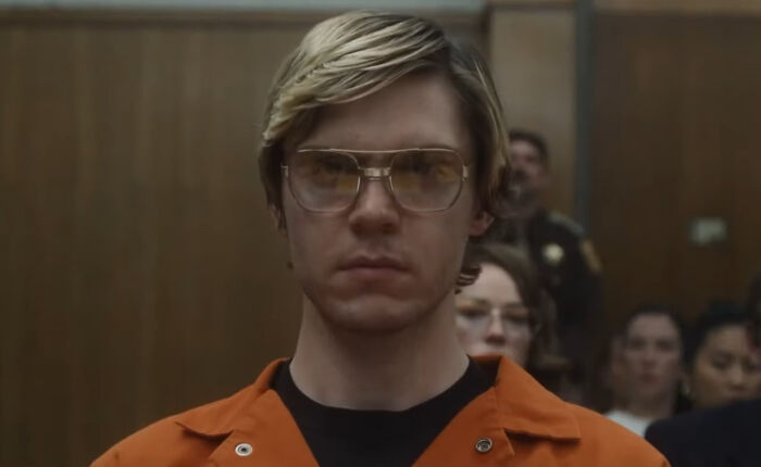 Young male actor in an orange prison jumpsuit wearing large glasses, portraying a difficult role in a courtroom scene.