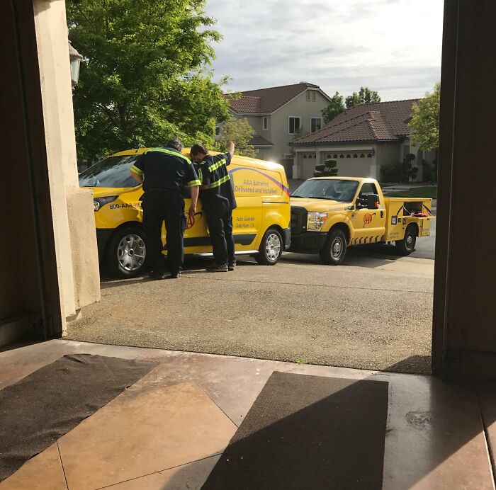 Two mechanics examining a broken-down AAA service van, parked in a residential driveway on a sunny day.