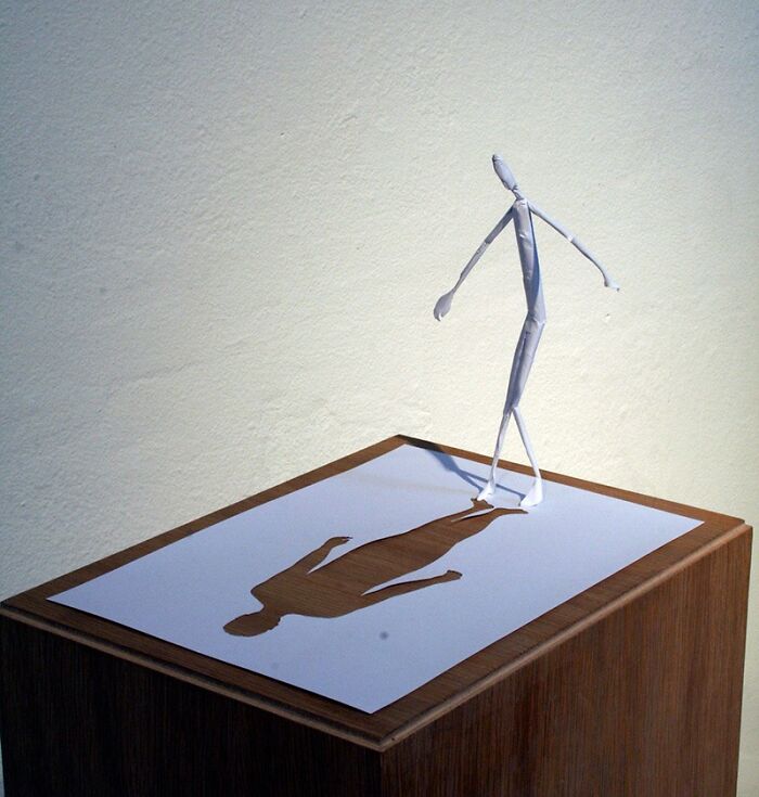 Mini world paper sculpture by Peter Callesen, featuring a cutout figure casting a shadow on a wooden pedestal.