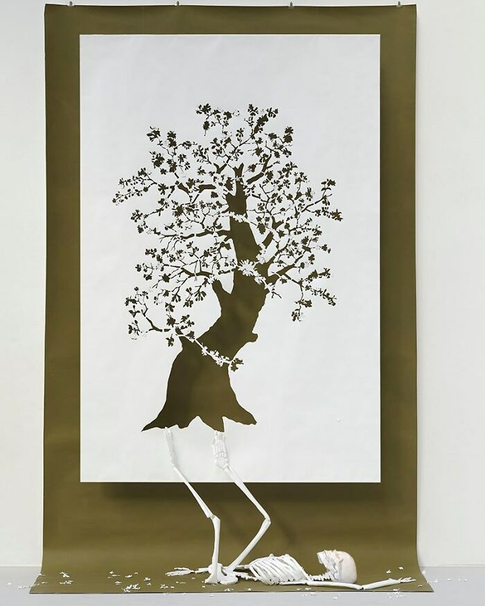 Paper art mini world depicting a tree with intricate cutouts and a skeleton below, crafted by Peter Callesen.