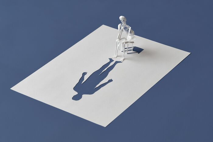Paper mini world featuring a skeleton on a chair casting a shadow on a white sheet, by Peter Callesen.