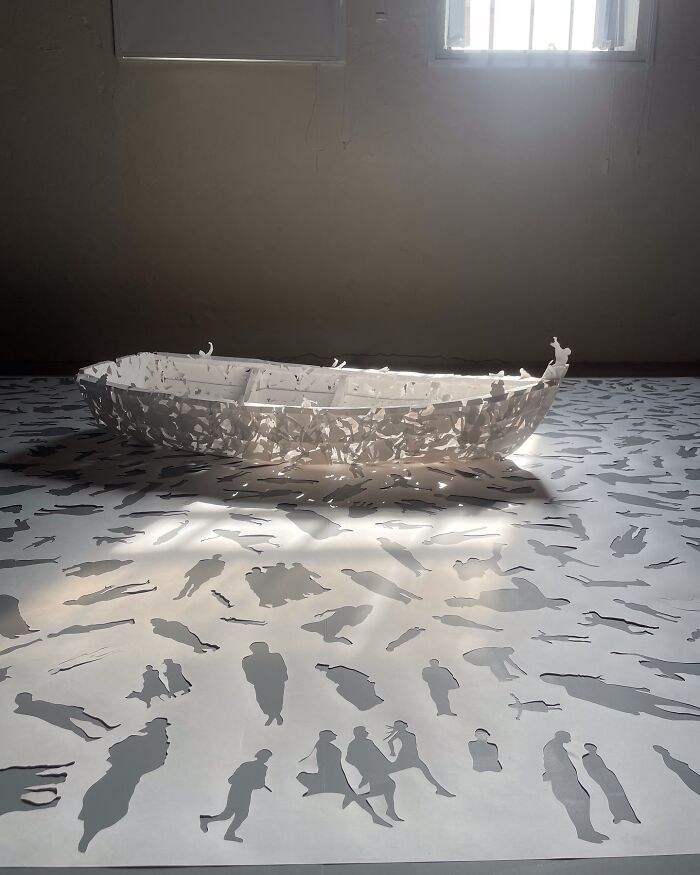 Paper art by Peter Callesen featuring a delicate boat, surrounded by intricate cut-out figures on a sunlit surface.