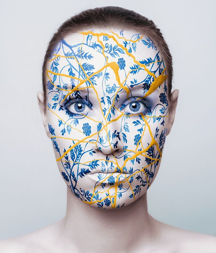 Surreal self-portrait of an artist with face painted in blue floral patterns and yellow lines.