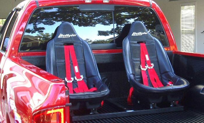 Two black BedRyder seats with red harnesses installed in the bed of a red pickup truck, Shark Tank products flop example.