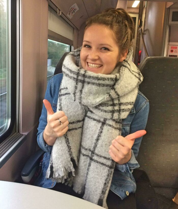 Smiling woman with a large plaid scarf giving thumbs up, capturing pure and uplifting moments on a train ride.
