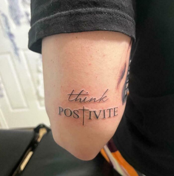 Tattoo fail on arm showing misspelled word positivity with a cross design replacing the letter T.