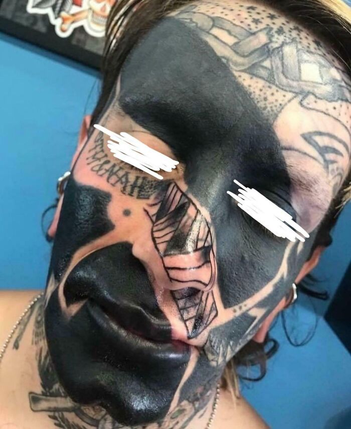 Extreme face tattoo fail covering most of the face in solid black ink with additional tattoo designs visible.