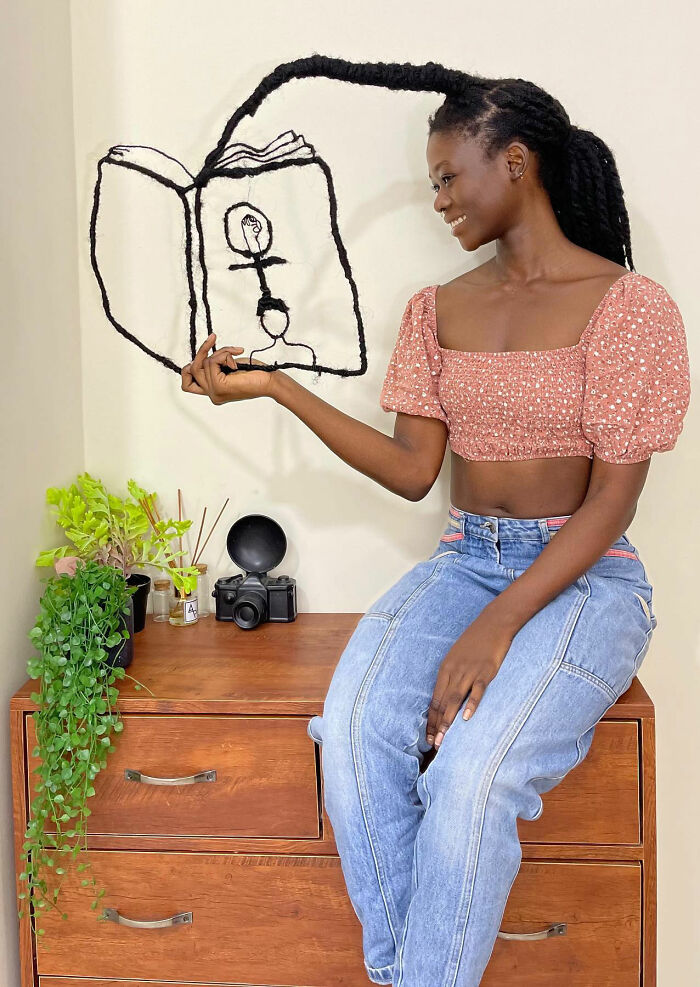 Artist with hair sculpture of a book, dressed in a floral top and jeans, seated on a dresser with plants and camera.