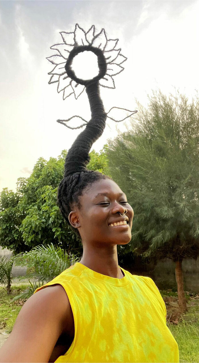 Artist with hair styled into a sunflower sculpture, wearing a yellow shirt, smiling outdoors.