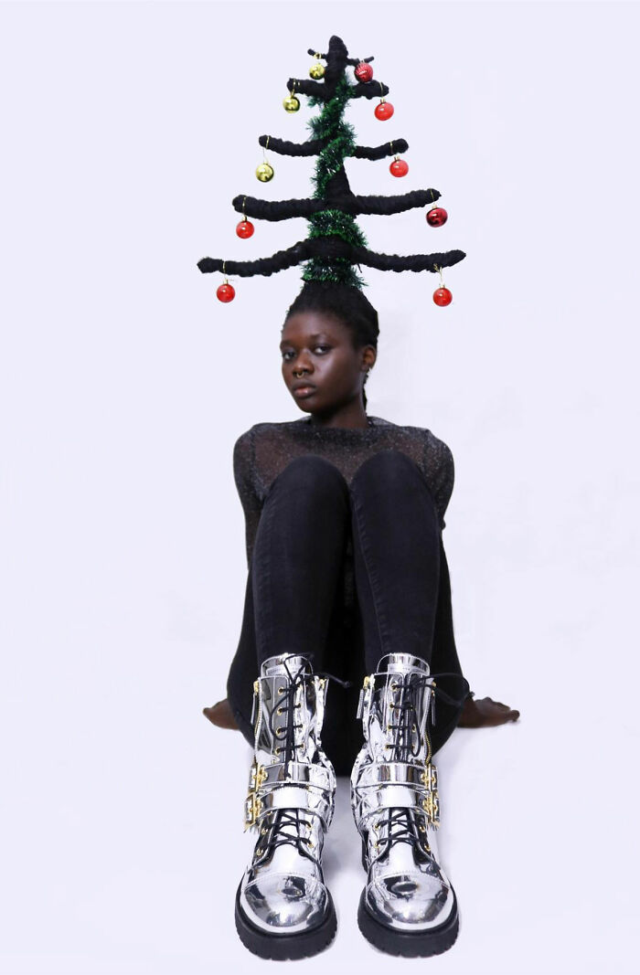 Artist with hair sculpture resembling a Christmas tree, adorned with ornaments, seated and wearing silver boots.