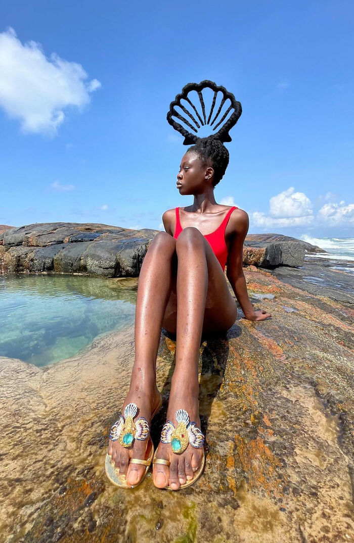 Artist with hair sculpture, sitting by the sea in a red swimsuit and decorative sandals.