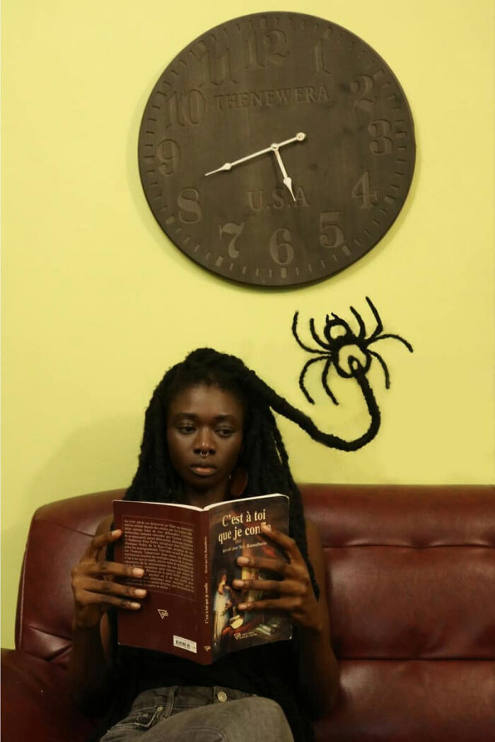 Artist with hair sculpted into a spider shape, reads a book under a large wall clock.
