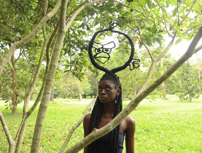 Artist with hair sculpture resembling an angry bird, standing in a lush green forest.