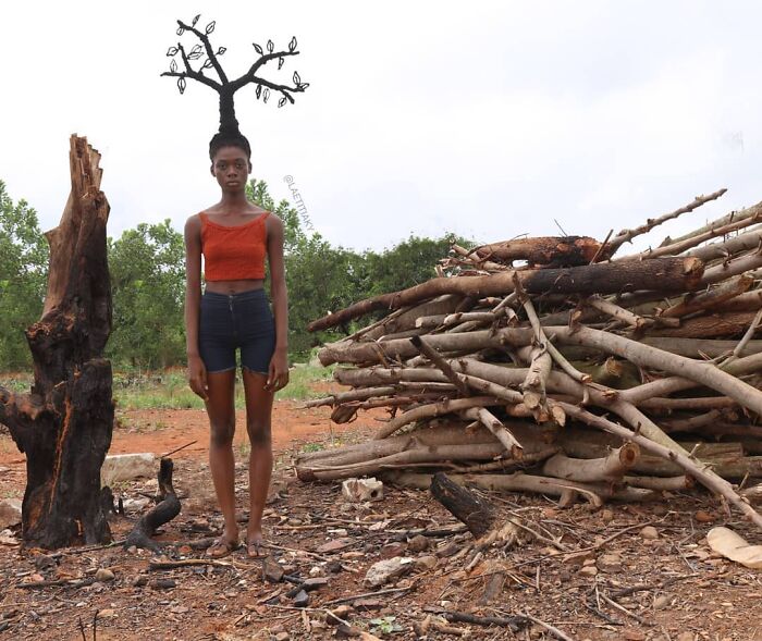 Artist with hair sculpture shaped like a tree, standing by logs in an outdoor setting.