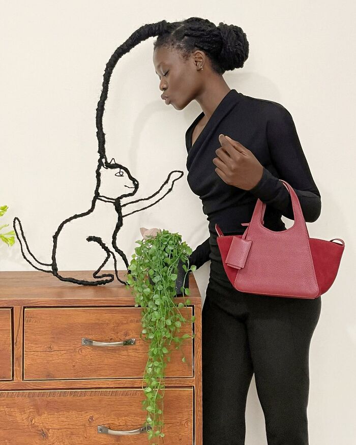 Artist with incredible hair sculpture resembling a cat, standing beside a wooden dresser, holding a red bag.