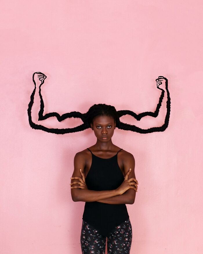 Artist with hair sculpted into muscular arms against a pink background, showcasing hair sculptures.